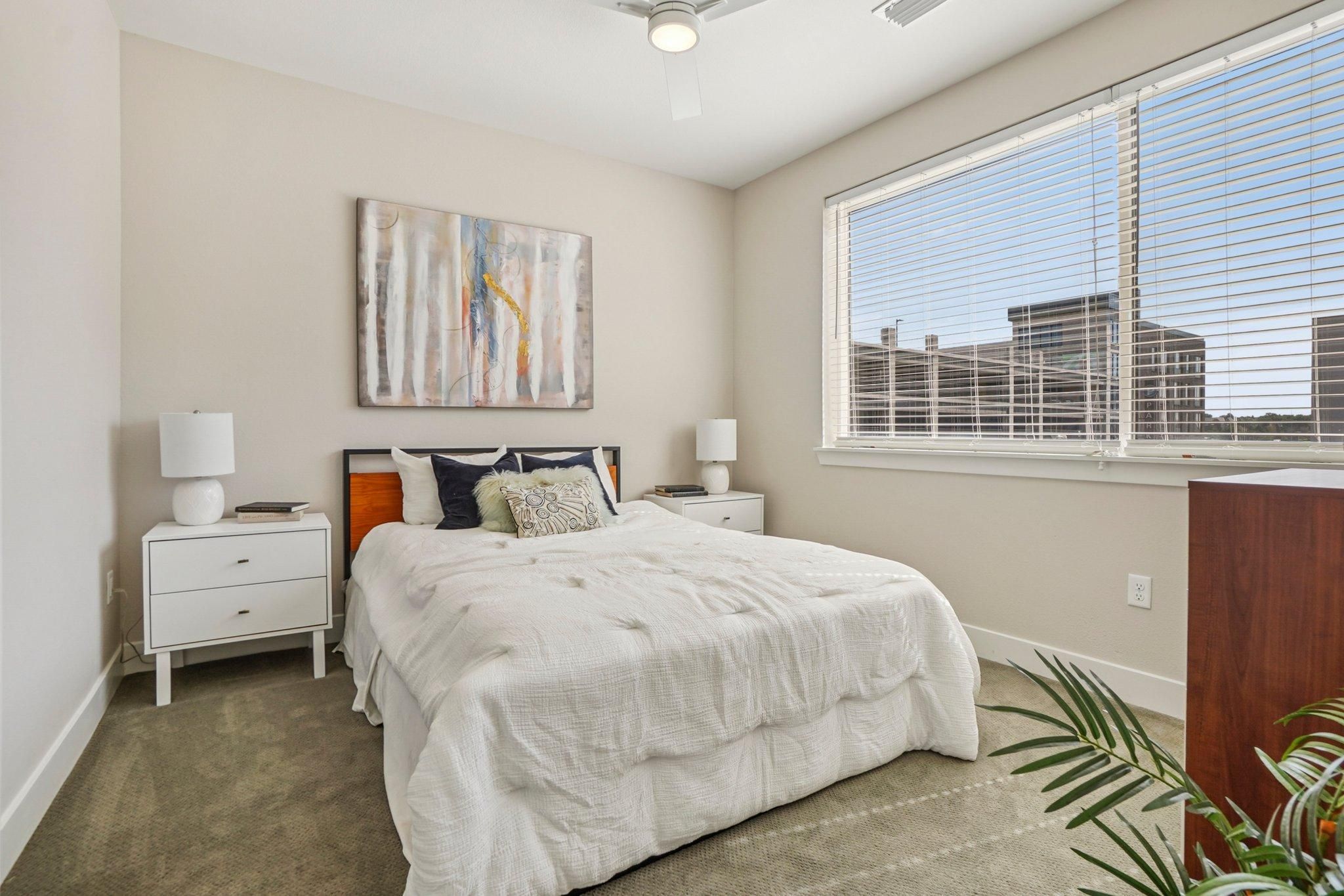 Ascent bright bedroom with a large window, bed with white bedding, two nightstands, and abstract wall art.